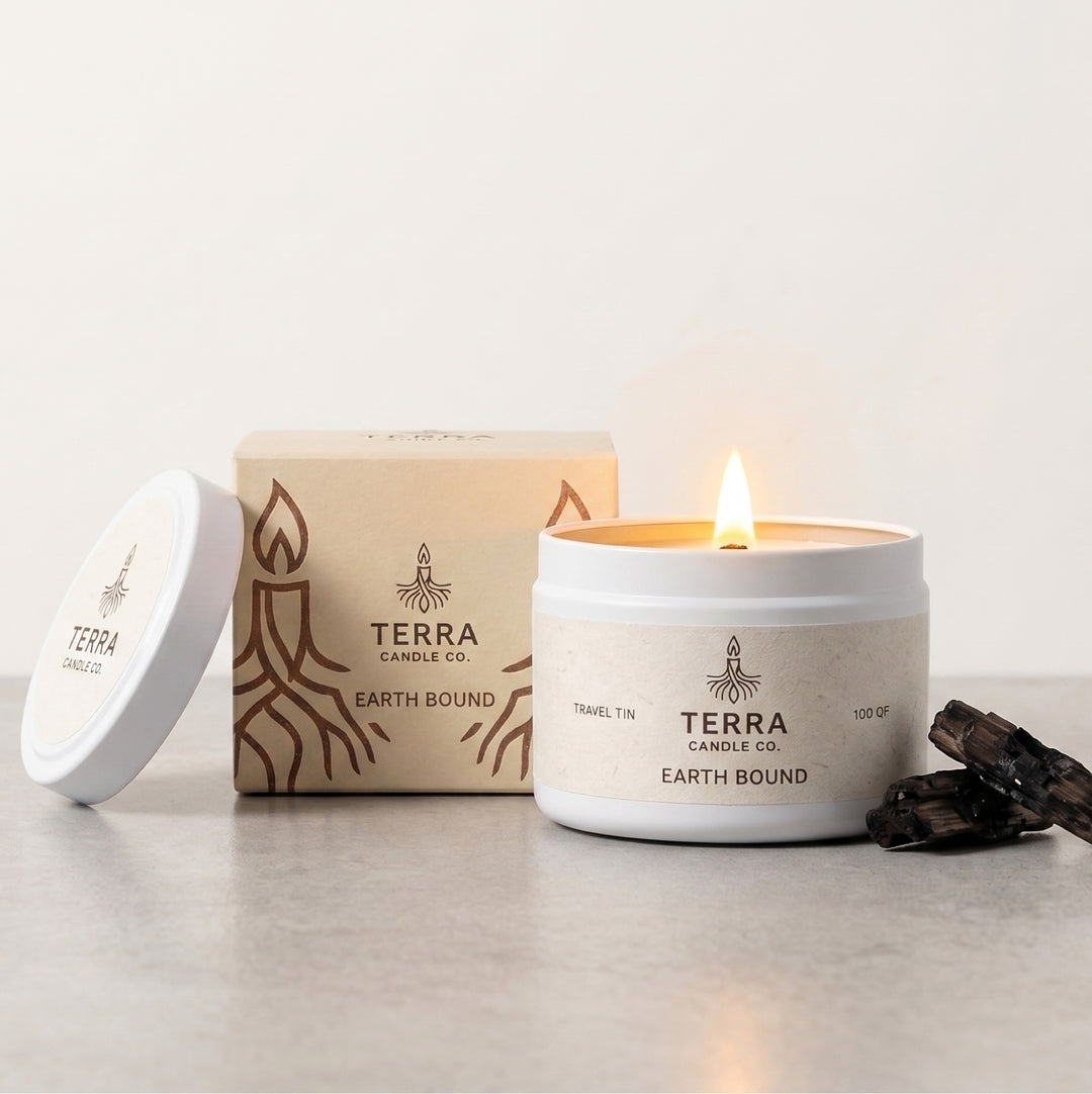 A Terra Candle Co. Earth Bound scented candle in a white jar with a lit flame, shown next to its beige and brown illustrated box and a white lid on a light gray surface.