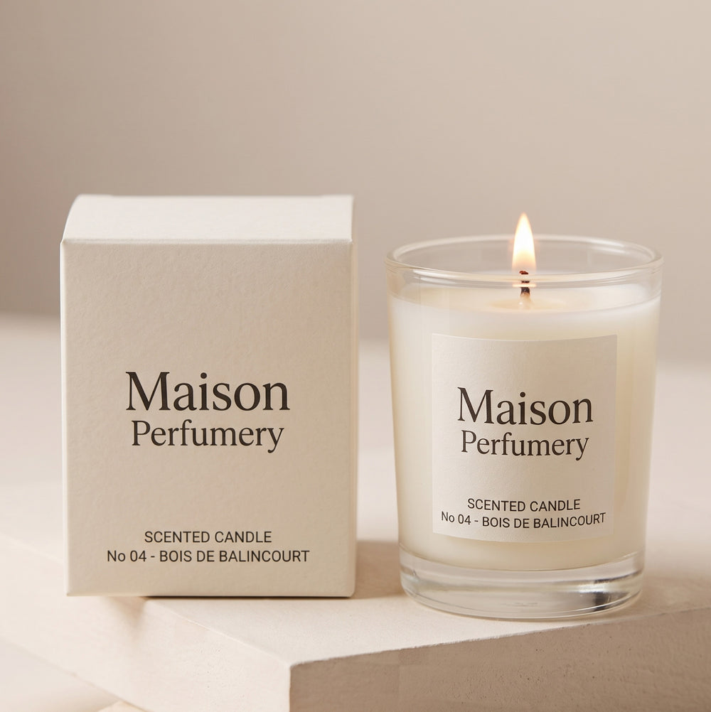 A Maison Perfumy scented candle in a clear glass jar with a lit flame, shown next to its cream-colored packaging box on a neutral beige background.