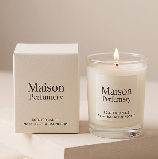 A Maison Perfumy scented candle in a clear glass jar with a lit flame, shown next to its cream-colored packaging box on a neutral beige background.