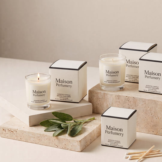 Maison Perfumy scented candles in clear glass jars with white labels, displayed alongside their white and black square boxes on stone risers. One candle is lit, and a few wooden matches lie nearby on a neutral beige surface.